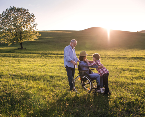 Rente bei Krankheit und Unfall: Großeltern und Enkel auf der Weide am spazieren. Das junge Mädchen hält die Oma auf dem Rollstuhl an den Schultern fest und lächelt Sie an.