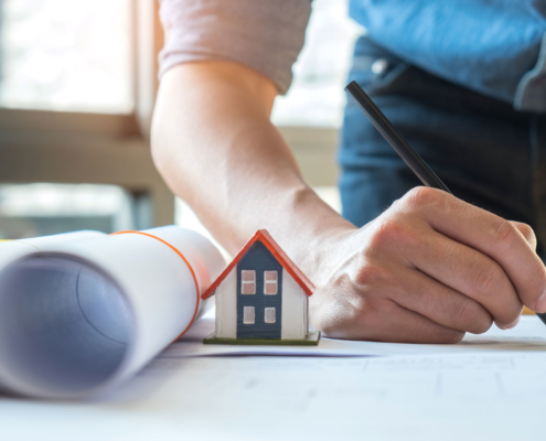Eigenheim Finanzierung: Ein Mann mit eingerolltem Hausplan und einem kleinen Haus auf dem Tischt unterschreibt gerade seine Eigenheim Finanzierung.