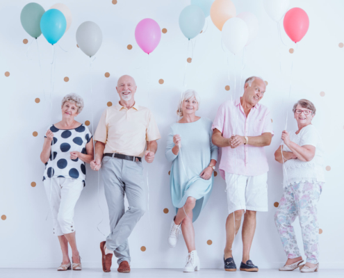 Pensionsplanung: Nach einer gut durchdachten Pensionsplanung erfolgt die Feier. Drei ältere Damen und zwei Herren posieren mit bunten Luftballons an einer weißen Wand und freuen sich über ihre Pensionierung.
