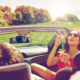 Fahrzeugversicherung: Mann fährt Auto. Eine Fahrzeugversicherung wurde abgeschlossen. Frau mit Sonnenbrille sitzt daneben und pustet Seifenblasen nach hinten zur Kamera.