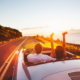 Oldtimerversicherung: Ein junges Paar fahren in einem Oldtimer durch eine Küstenstraße beim Sonnenuntergang.